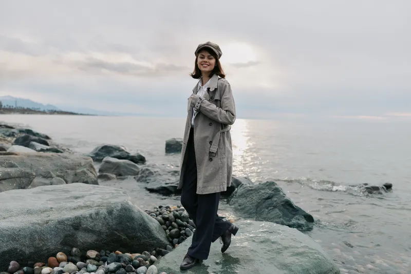 Young woman in plaid coat and hat stands on rocky shore near calm sea with cloudy sky. Casual autumn fashion concept with nature and outdoor lifestyle background.