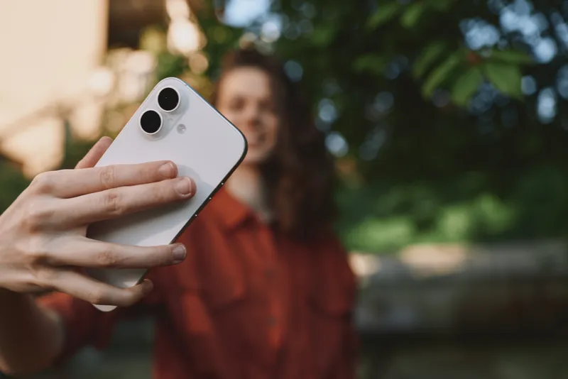 A casual woman outdoors taking a selfie with a smartphone, wearing a red shirt and smiling. Bright greenery surrounds her, capturing everyday life and self expression