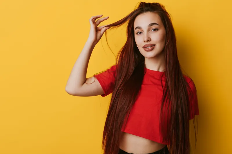 Young woman with long hair wearing red crop top posing on yellow background. Female model smiling with hand in hair for beauty and fashion portrait studio shot.