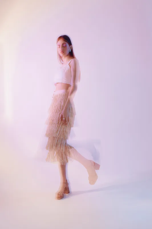 Young woman in stylish beige outfit poses with one leg raised on soft pink background. Concept of woman beauty fashion, elegance, and modern style in studio lighting.