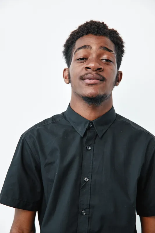 Young black man with dark skin wearing black shirt standing against white background looking confident and relaxed with slight smile and short curly hair in studio portrait.