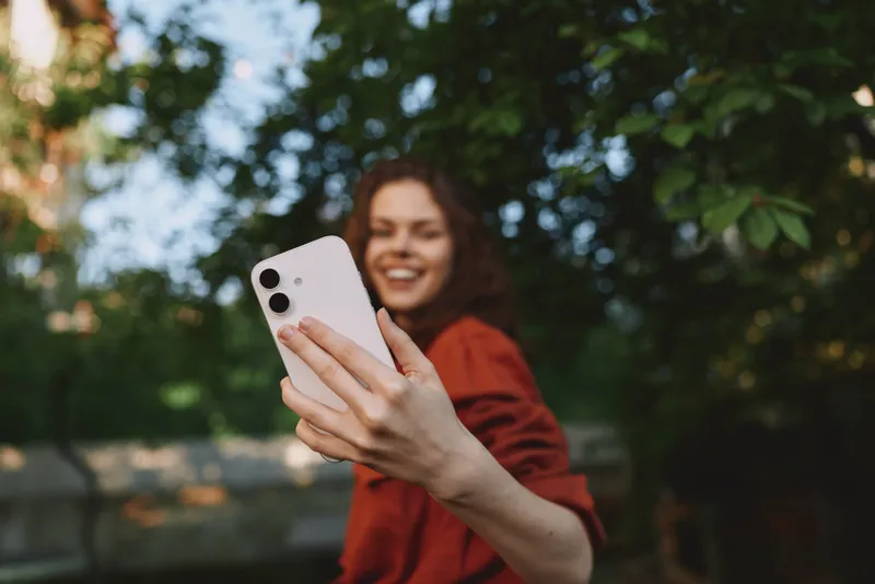 Young woman with curly hair takes a cheerful selfie outdoors, extending a smartphone toward the camera while smiling under bright daylight with green trees in the background