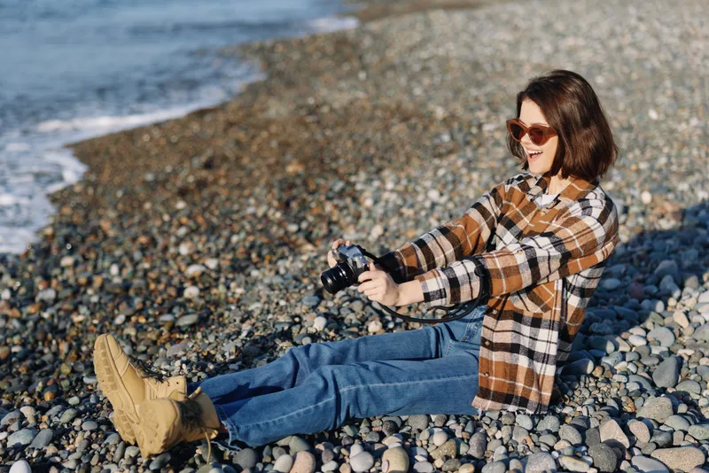 Woman sits on a rocky beach in a plaid shirt and jeans, capturing a candid moment with a compact camera as waves roll in and sunlight glints off the shore.