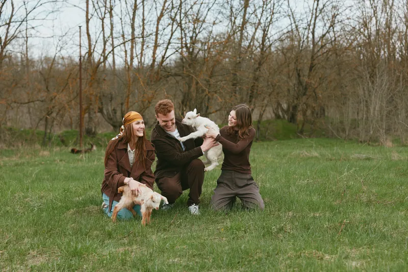 Three young adults enjoy a playful moment with two baby goats in a grassy field surrounded by bare trees during early spring outdoors.