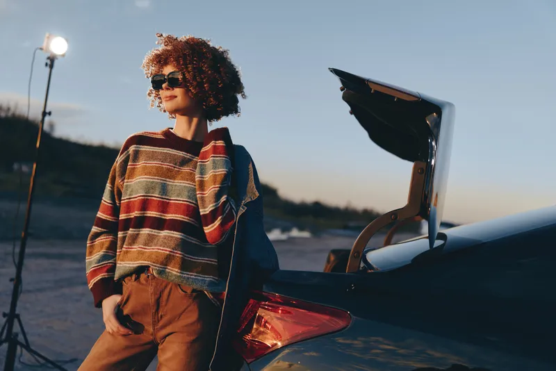 Woman with curly hair stands outdoors by open car trunk at sunset, wearing sunglasses and a patterned sweater, graduation cap near the vehicle, woman lifestyle
