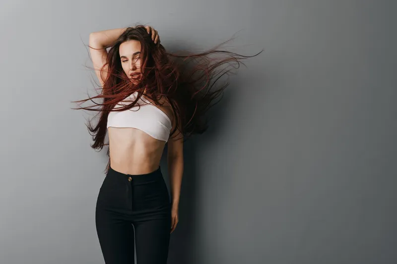 Woman with long hair posing in white crop top and black pants against gray background. Fashion, beauty and confident model with casual style and dynamic hair movement indoors.