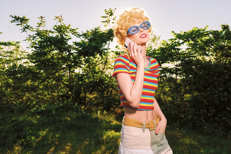 Smiling woman with curly blonde hair wearing a striped crop top stands outdoors on a sunny day, exuding retro fashion and lifestyle. Natural light enhances color, pose, and a cheerful mood.