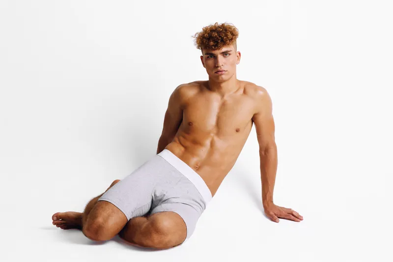 Young man with curly hair sitting on floor in gray shorts against white background, showing muscular torso and relaxed pose for fitness or lifestyle concept.