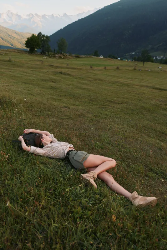 A young woman in casual clothes lies relaxed on green grass in a mountain valley, enjoying nature and peaceful outdoor lifestyle during a sunny day.
