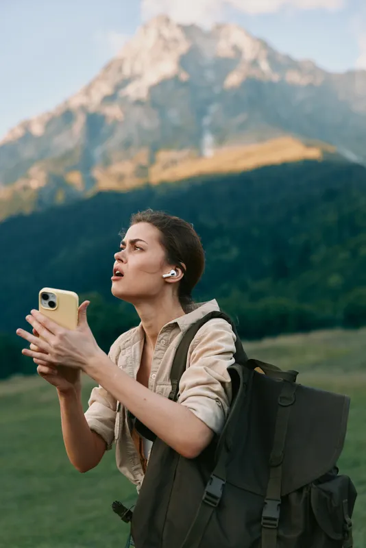 A young woman with a backpack outdoors holds a smartphone and looks concerned while standing in a mountain landscape. Concept for travel, communication, and lifestyle.