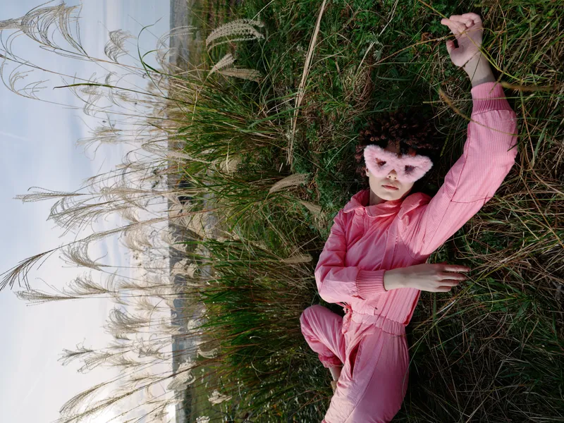 Person in a pink suit wearing an animal mask lying in tall grass in an open field, exploring nature with a playful, mysterious mood in a dramatic outdoor portrait