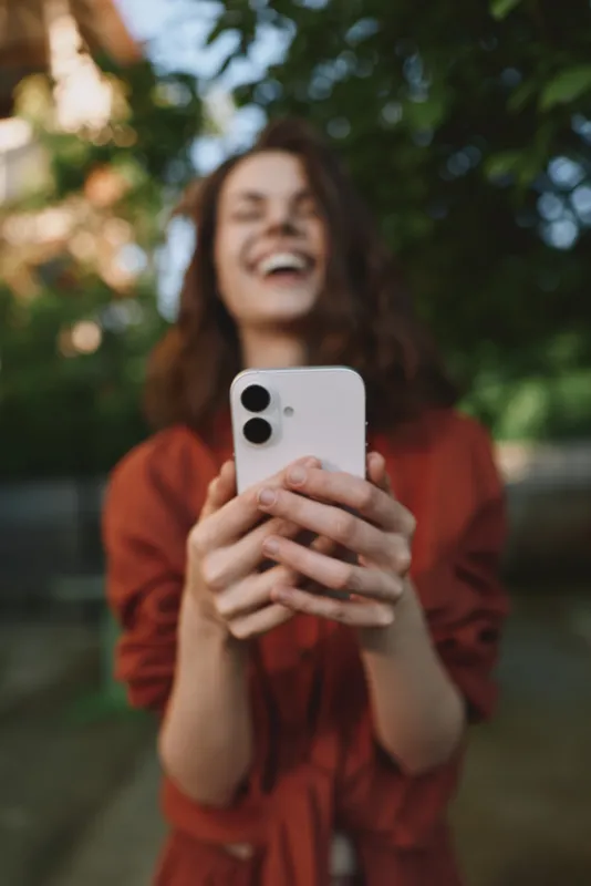 Young woman outdoors laughs while taking a selfie with a smartphone, wearing a rust top, sunlit leaves in the background, relaxed pose, natural colors, candid moment