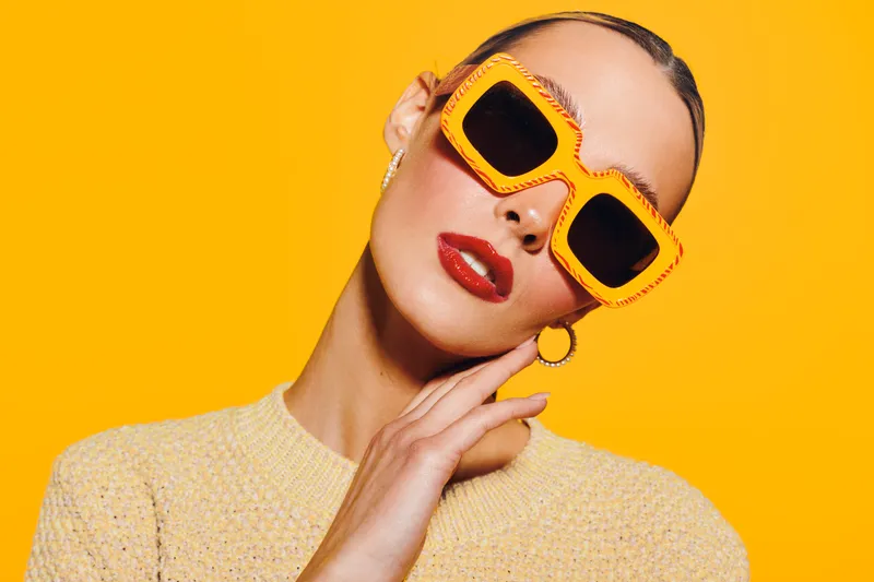 Stylish woman with slicked hair wearing large orange sunglasses and red lipstick poses against bright yellow background. Fashion concept with bold colors and modern accessories.