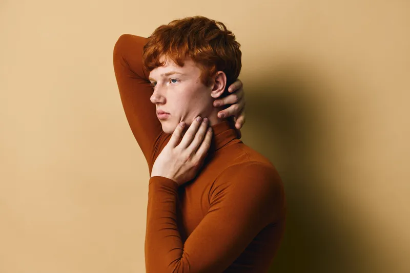 Young man with red hair posing with hand on neck in brown turtleneck sweater against beige background. Side profile portrait of serious confident male model in soft studio light.