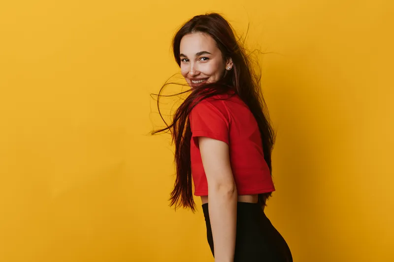 Young woman with long hair wearing red top and black pants posing and smiling against yellow background. Female model with casual style and natural expression in studio portrait.