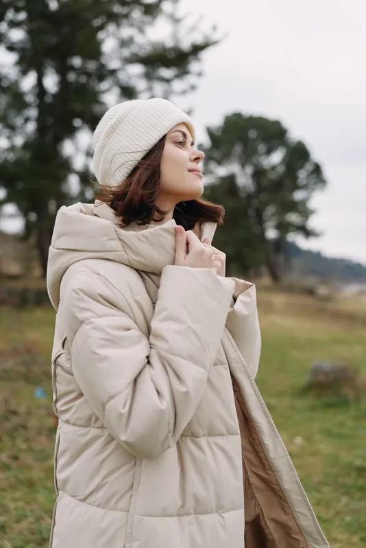 woman outdoor portrait in winter coat, cream beanie, soft light, wind lifts her jacket as she stands in a calm countryside field, showcasing cozy fashion and natural texture