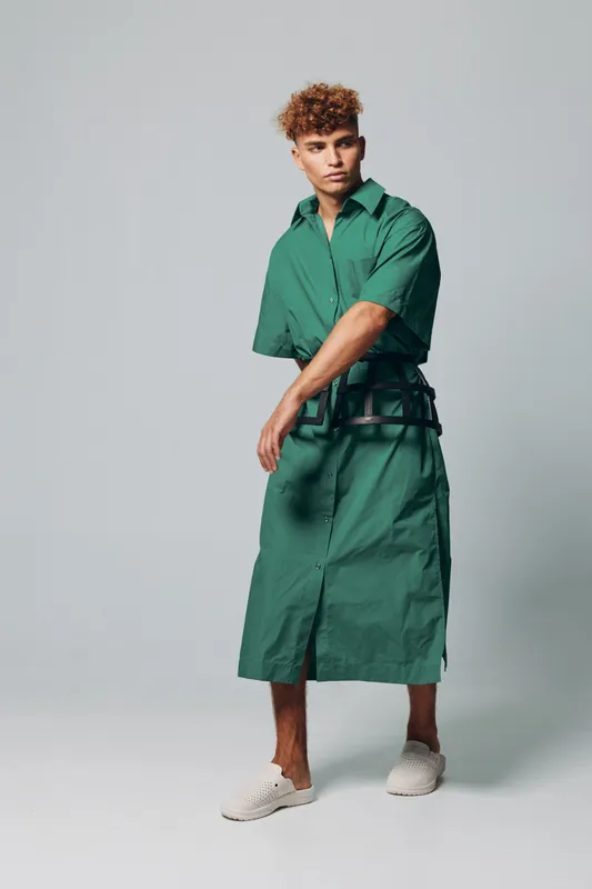 Young man with curly hair wearing green long shirt dress and belt posing in studio with neutral background. Casual fashion style with white slip-on shoes and confident expression.