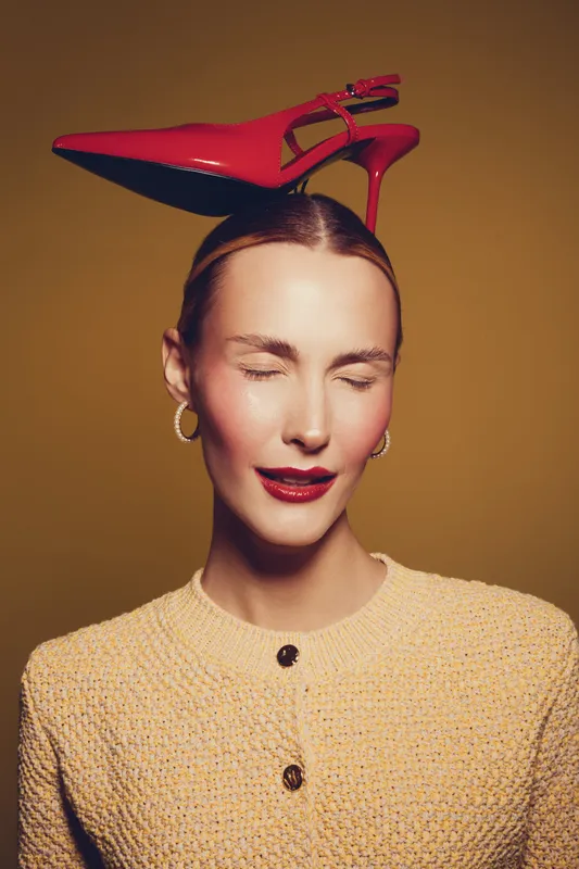 Young woman with closed eyes wearing a beige textured jacket and red high heel shoe balanced on her head. Studio portrait with warm lighting and fashion concept.