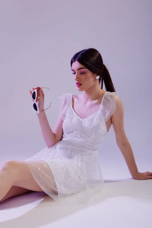Young woman in a white lace dress holds sunglasses while sitting on the floor. Soft lighting highlights her beauty and fashion style in a minimal studio setting. Concept for elegance.