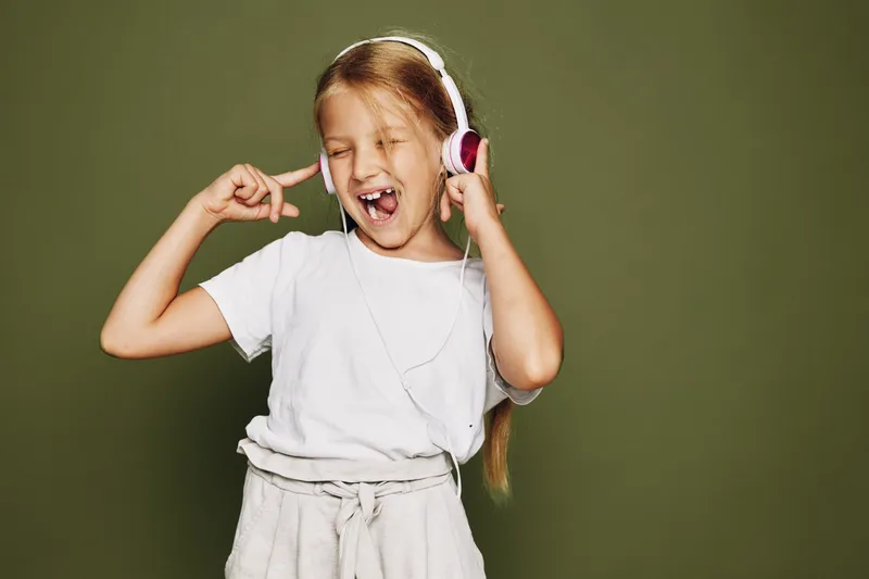 Happy girl in white t-shirt wearing headphones and singing with eyes closed against green background. Concept for music, fun, and childhood joy.