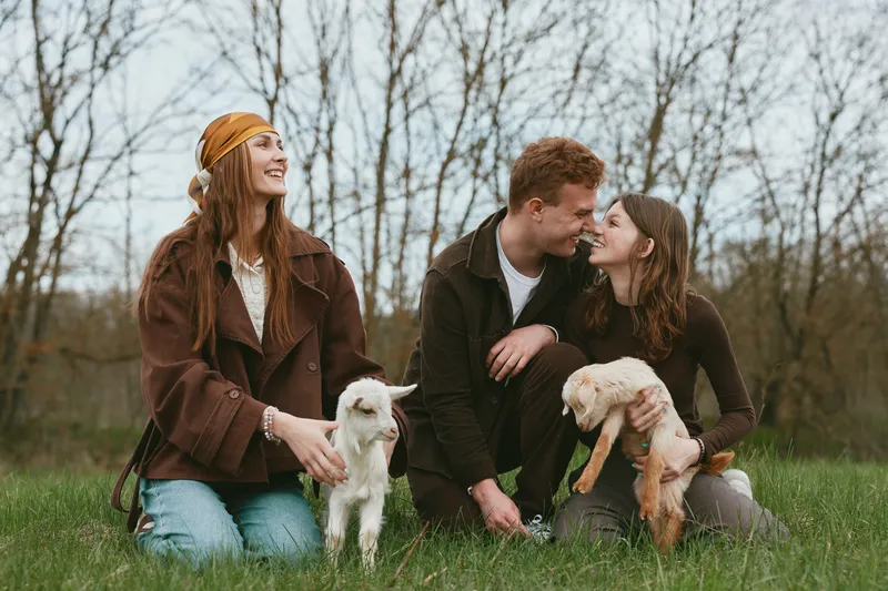 Young couple and woman enjoy time outdoors with two baby goats in a grassy field during early spring, sharing joyful moments and natural connection in casual warm clothing.