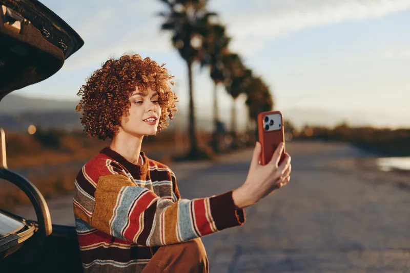 Woman lifestyle outdoor, selfie with smartphone, curly hair, striped sweater, by a parked car on a street at golden hour, warm relaxed lifestyle