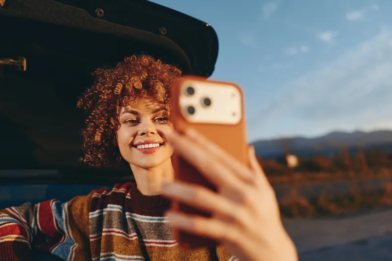 Woman outdoors beside a car, smiling for a selfie with smartphone, curly hair catching warm sunset light, casual lifestyle moment, mountains and sky softly blurred, natural colors.
