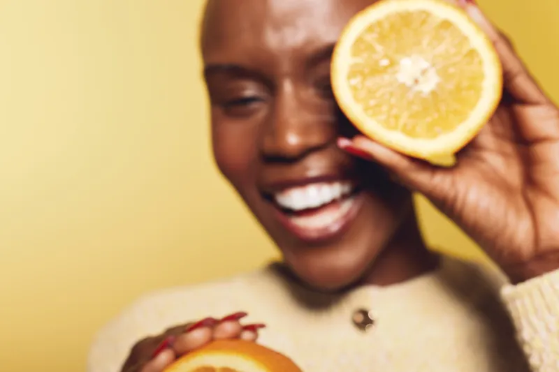 Happy African woman with dark skin holding orange slices near face, smiling with closed eyes on yellow background. Concept of healthy lifestyle, vitamin C, and natural beauty.