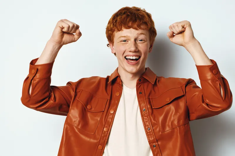 Young man with red hair wearing brown shirt smiling and raising fists with excitement against white background, cheerful male portrait, casual style, happy emotion, positive gesture