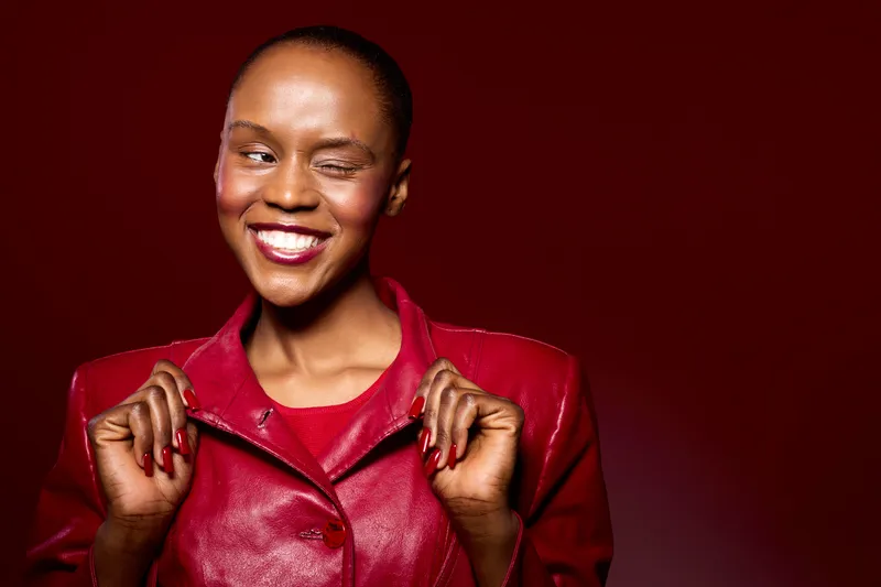 Confident black woman with dark skin winks and smiles wearing a red leather jacket against a red background. Concept of fashion, beauty, and self-expression with vibrant colors.