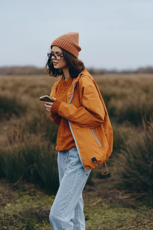 Woman outdoors in field autumn, walking in profile, wearing orange jacket and knitted beanie, crossbody bag, casual jeans, lifestyle concept, nature calm, soft sunset light