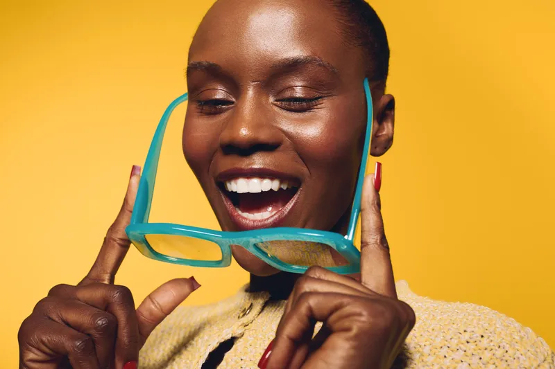 Happy African woman with dark skin holds bright blue sunglasses near face, smiling with closed eyes against yellow background. Concept of joy, fashion, and summer style with vibrant colors.