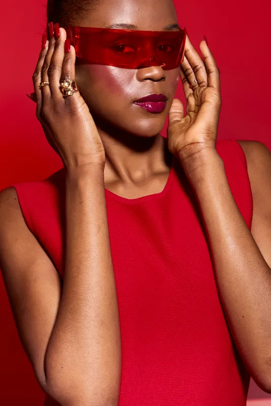 Stylish dark skinned African woman wearing red dress and red translucent visor poses on red background. Fashion portrait with glossy makeup, rings, and confident expression.