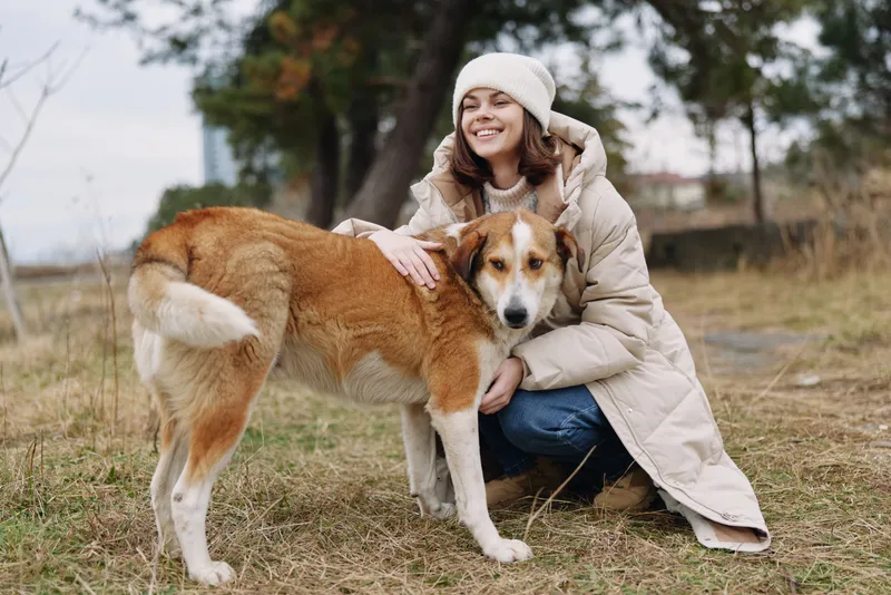 Woman in a warm coat crouches beside a friendly dog in an open park, sharing a gentle hug as autumn grass, colorful leaves and tall trees frame the peaceful outdoor moment for a tender, candid