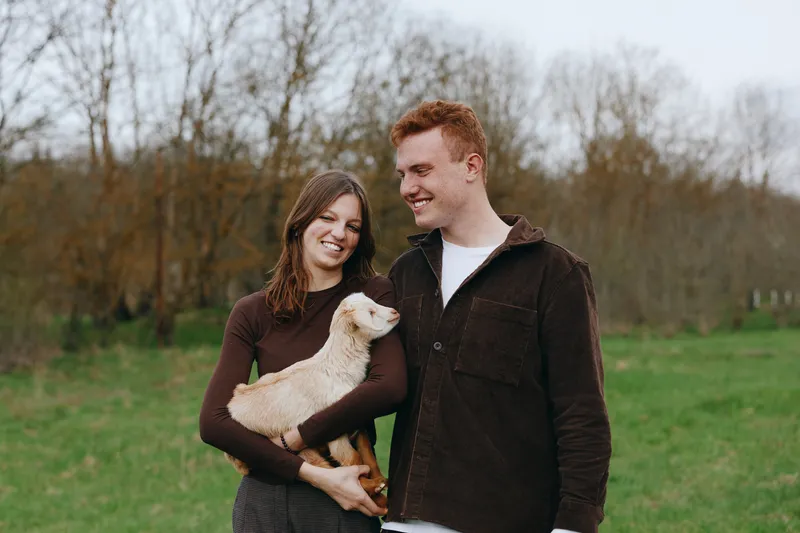 Young couple enjoys a moment outdoors as the woman holds a small baby goat in her arms while both smile in a natural countryside setting during autumn.