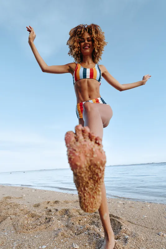 Woman in striped swimsuit jumps on sandy beach, curly hair flying in sea breeze, joyful summer vacation mood under clear blue sky, seaside leisure and freedom