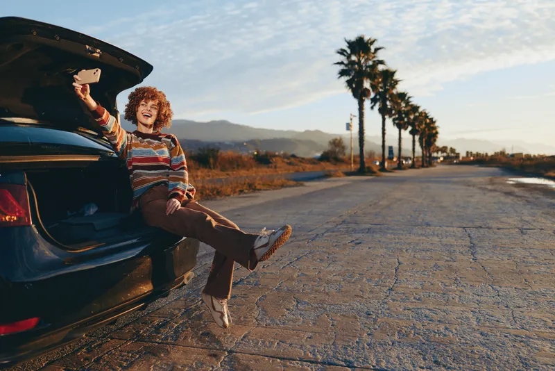 Woman lifestyle outdoors, seated beside open car door on road, casual striped sweater and skirt, relaxed walking pose, warm sunset light, palm trees line avenue, hills in distance