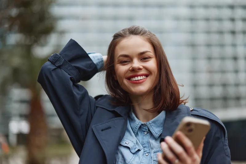 Young woman with brunette hair smiles happily while holding a smartphone outdoors in casual denim shirt and dark jacket during daytime.