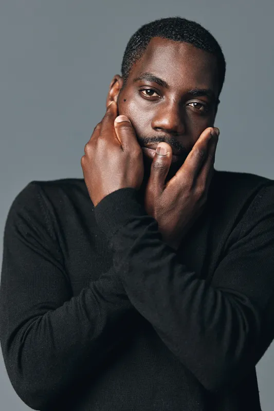 African man with dark skin wearing black sweater posing with hands on face in creative conceptual visual style. Studio portrait with serious expression and artistic mood on gray background.