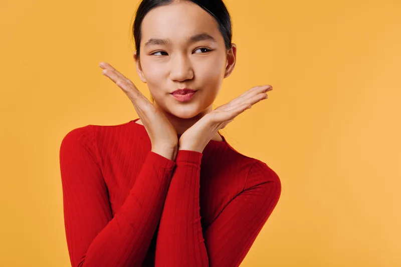 Woman in red sweater posing on orange background with hands near face, smiling and looking to side with playful expression and casual style in studio portrait photography.