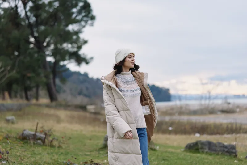 Woman in a warm coat and hat enjoying outdoor scenery by a lake, embracing winter fashion amid natural landscape with trees and calm breeze