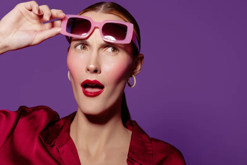 Surprised woman with pink sunglasses and red lipstick looks away on purple background. Fashion concept with expressive face and bright colors in studio lighting.