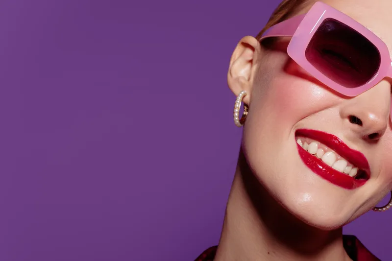 Close-up of smiling woman with red lipstick and pink sunglasses on purple background. Fashion portrait with glossy skin, hoop earrings, and confident expression. Concept of style and beauty.