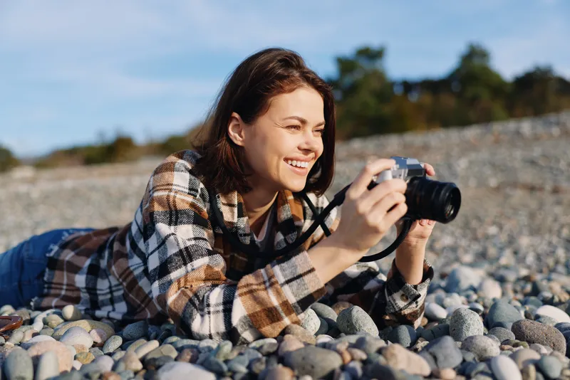 Woman photographer lying on a pebble beach with a camera, smiling as she captures scenery. Casual plaid shirt, outdoor light, relaxed creative moment by the sea and stones