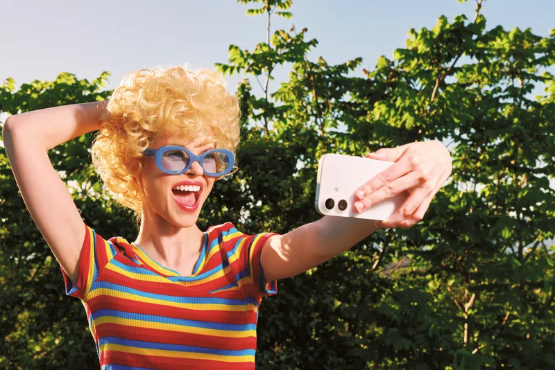 Smiling woman with curly blonde hair wearing a striped shirt and blue sunglasses takes a cheerful selfie outdoors on a sunny day. Colorful retro vibes enhance lifestyle and happiness.
