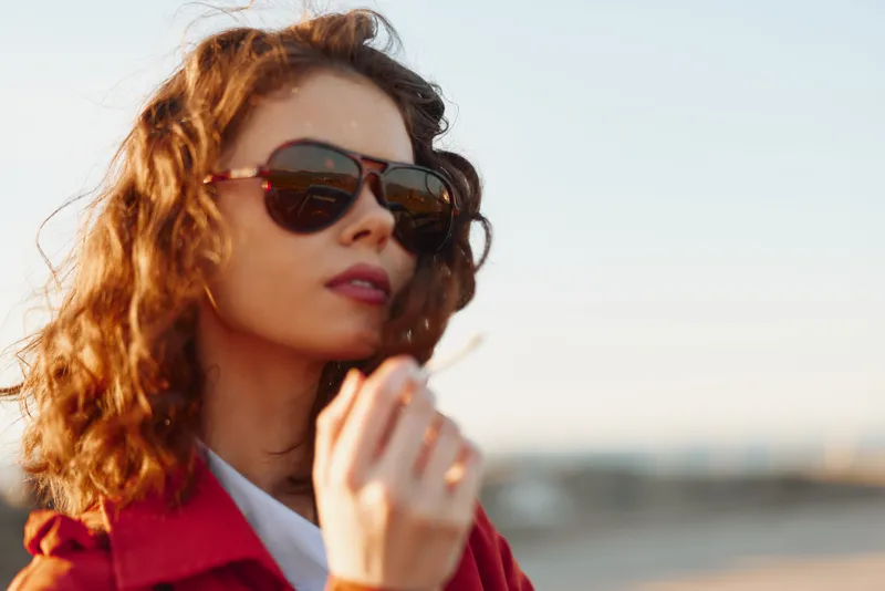 Young woman with curly hair wearing sunglasses stands outdoors, looking away with a confident expression in warm light, red jacket framing her silhouette against a clear sky.