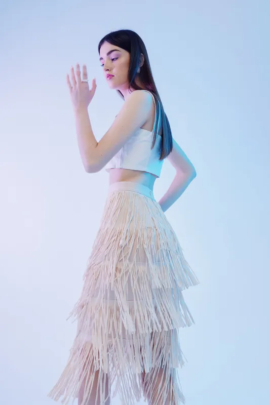 Young woman in a white crop top and layered fringe skirt poses with closed eyes in a soft studio light. Concept of fashion, beauty, and modern style.
