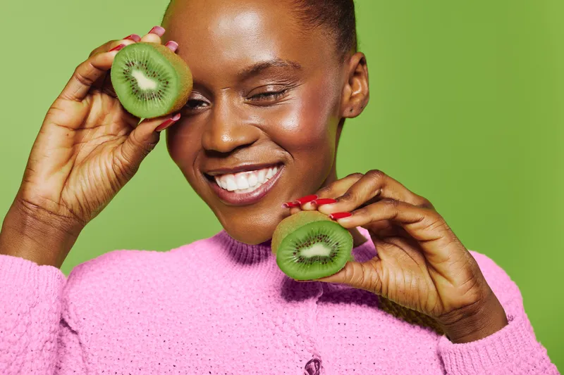 Smiling black woman with dark skin holds two kiwi halves near her face wearing pink sweater on green background. Concept of healthy eating, natural beauty, and fresh fruit lifestyle.