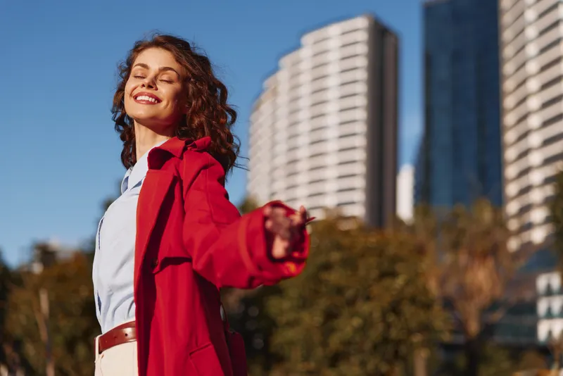 Young woman with curly hair in a red coat and white shirt smiles with eyes closed in bright sunlight, posing outdoors with modern buildings in the background, feeling free and joyful.