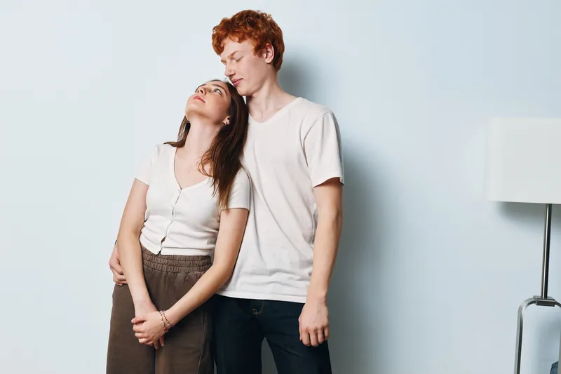 Young couple embracing indoors with woman leaning on man showing affection and comfort. Casual clothing, standing against plain wall in cozy intimate moment between lovers.